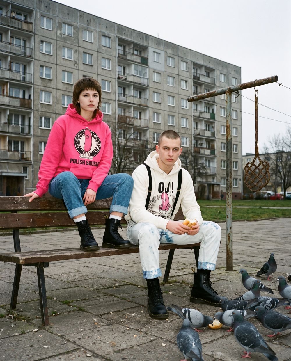 Dziewczyna w różowej bluzie 0 proc. patrioty i chłopak w bluzie Oj! na ławce pod blokiem, karmią gołębie